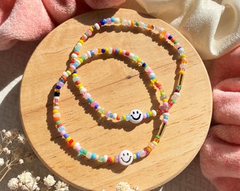 May include: Two beaded bracelets with a rainbow of colors. Each bracelet features a white smiley face bead. The bracelets are on a wooden surface, with pink and white fabric in the background. The bracelets are a fun and colorful accessory.