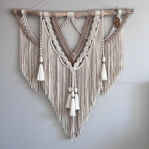 May include: A beige and white macrame wall hanging with a wooden dowel at the top. The macrame is made with a variety of knots and has a fringe at the bottom. There are three tassels at the bottom of the hanging.