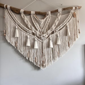 May include: A large macrame wall hanging with a natural wood branch at the top. The macrame is made with cream-coloured cord and features a variety of knots and patterns. The hanging is decorated with small wooden beads.