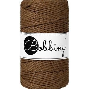 Op de afbeelding: Een strak opgerolde spoel bruin macramé koord. Een wit etiket met het woord "Bobbiny" in een scriptlettertype is om het midden van de spoel gewikkeld. Het koord heeft een effen, aardse bruine kleur.