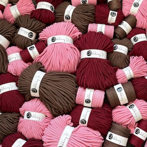 Op de afbeelding: Een verzameling van meerdere strengen Bobbiny katoenkoord in diverse tinten roze, donkerrood en bruin. Elke streng is omwikkeld met een wit papieren label met het Bobbiny merklogo en tekst, gerangschikt in een dichte, overlappende stapel.