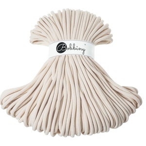 May include: A close-up of a spool of natural colored cotton cord. The cord is wrapped around itself in a circular shape. The spool is labeled "Bobbiny".