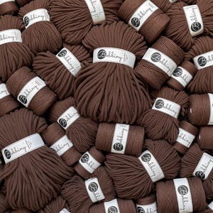 Può includere: Una collezione di diverse matasse di corda per macramè marrone scuro, ognuna avvolta da un'etichetta di carta bianca con il marchio Bobbiny e la scritta 5mm. La corda è spessa, intrecciata e disposta in un mucchio denso e sovrapposto.