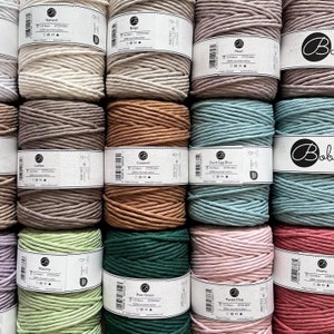 Op de afbeelding: Een verzameling spoelen kleurrijk macramé-garen in diverse tinten, waaronder beige, bruin, groen en roze. Elke spoel is gelabeld met de kleurnaam en bevat 112 meter garen van 100% gerecycled katoen. De garens zijn netjes opgestapeld en tonen een assortiment knutselbenodigdheden.