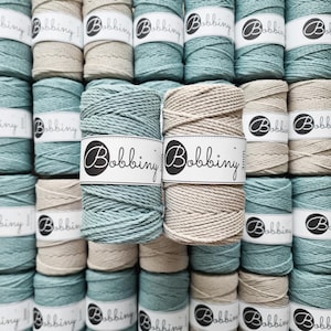 Può includere: Una griglia di rocchetti di filo per macramè in vari colori. I rocchetti sono etichettati "Bobbiny" e sono in tonalità di verde acqua e beige. Il filo sembra avere uno spessore di 3 mm.