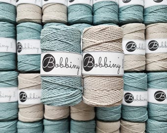 Könnte beinhalten: Eine Anordnung von Makramee-Garnrollen in verschiedenen Farben. Die Rollen sind mit "Bobbiny" beschriftet und in Türkis- und Beigetönen gehalten. Das Garn scheint 3 mm dick zu sein.