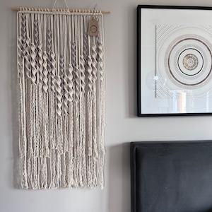 Puede incluir: Un tapiz de macramé beige y marrón con una barra de madera en la parte superior. El tapiz está hecho de cuerdas anudadas y tiene flecos en la parte inferior.