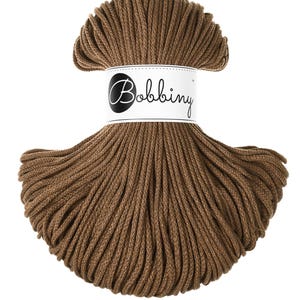 Può includere: Un gomitolo di filo marrone Bobbing per macramè. Il filo è avvolto strettamente e avvolto con un'etichetta bianca con il nome del marchio in un carattere nero. Il filo è di un colore marrone caldo e terroso.