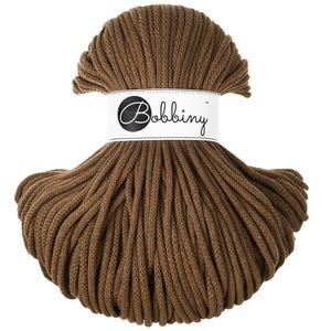 Puede incluir: Un carrete de hilo marrón de la marca Bobbiny. El cordón está bien enrollado y sujeto con una etiqueta blanca que dice "Bobbiny" en cursiva. El cordón es de color marrón tierra.