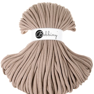 Könnte beinhalten: Nahaufnahme einer großen Spule mit beigefarbenem Baumwollgarn. Das Garn ist fest gewickelt und hat eine glatte Textur. Die Spule ist mit "Bobbiny" beschriftet.