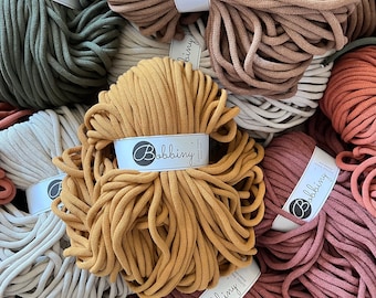Può includere: Una varietà di corde di cotone colorate, note anche come cordoncino per macramè, in diversi colori e trame. La corda è etichettata con il nome del marchio "Bobbiny".