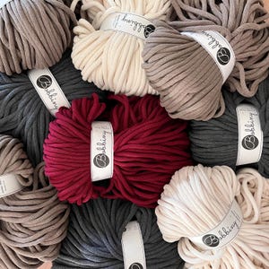 Peut inclure: Une collection de huit bobines de corde en coton épaisse de différentes couleurs, dont le beige, le brun, le gris, le rouge et le blanc. Chaque bobine porte une étiquette blanche avec le mot "Bobbiny" imprimé dessus.