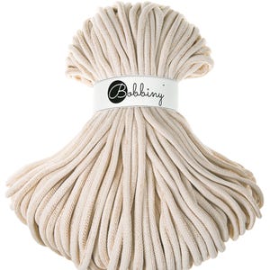 Puede incluir: Primer plano de una gran bobina de cordón de algodón de color crema. El cordón está enrollado apretado y tiene una superficie ligeramente texturizada. La bobina está etiquetada con "Bobbiny".