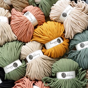 Puede incluir: Una variedad de cuerda de algodón en diferentes colores, incluyendo beige, verde, negro, naranja y amarillo. La cuerda está dispuesta en una pila y tiene una etiqueta que dice "Bobbiny".