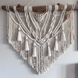 May include: A large, white macrame wall hanging with a wooden branch at the top. The macrame features a geometric design with tassels and wooden beads.