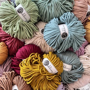 May include: A variety of colorful cotton rope, also known as macrame cord, in different colors, including green, blue, red, yellow, and brown. The rope is arranged in a pile and each spool has a white label with the word "Bobbiny" printed on it.