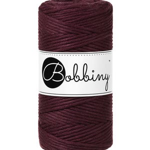 Op de afbeelding: Een spoel bordeauxkleurig macramé-garen. Het garen is strak gewikkeld en om een cilindrische kern gewikkeld. Een wit etiket met het woord "Bobbiny" in een scriptlettertype is rond het midden van de spoel gewikkeld.