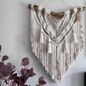 Op de afbeelding: Een witte macramé wandhanger met een houten tak aan de bovenkant en houten kralen die er doorheen geweven zijn. De macramé heeft de vorm van een hart en heeft een franje aan de onderkant.