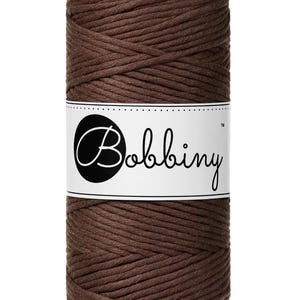 May include: A cylindrical spool of dark brown cotton cord wrapped in a tight, continuous coil. A white paper label is wrapped around the center of the spool, featuring the word Bobbiny printed in a black, cursive script inside a black circle.