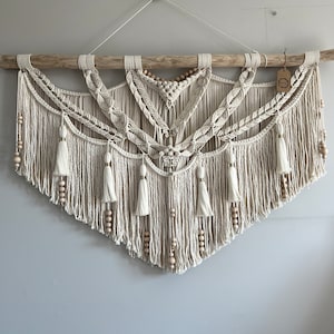 以下が含まれることがあります： 流木をベースにした、クリーム色のマクラメの壁掛け。複雑な結び目、編み込み部分、吊り下げられたタッセルが特徴です。木製のビーズが組み込まれています。右側にタグが付いています。壁の装飾。
