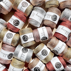 Può includere: Una collezione di bobine di cordoncino testurizzato da 3 mm di spessore, dai colori vivaci, con il nome del marchio "Bobbiny" stampato su ogni bobina. Le bobine sono disposte in modo casuale su una superficie di legno.