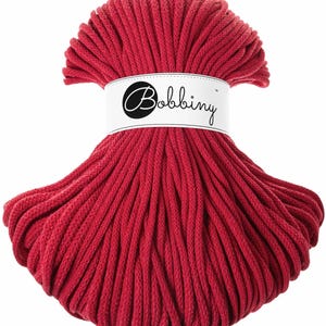 May include: A bundle of red macrame cord with a white label wrapped around the centre. The label features the word "Bobbiny" in a script font. The cord is tightly wound and appears soft, suitable for various crafting projects.