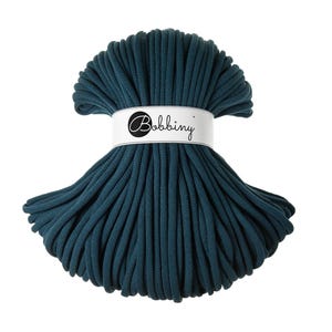 Può includere: Un gomitolo di cordoncino per macramè color teal. Il cordoncino è avvolto strettamente e tenuto insieme da una fascia bianca con la scritta "Bobbiny" in nero. Il cordoncino è di un colore teal intenso.