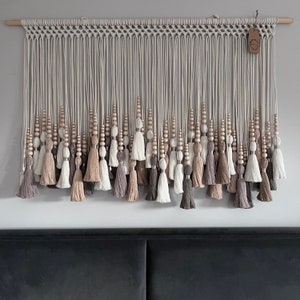 Puede incluir: Un tapiz de macramé para colgar en la pared con un patrón tejido y una franja de borlas en tonos beige, marrón y blanco.