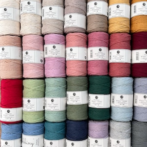 Op de afbeelding: Een verzameling kleurrijke macramé-garenspoelen in verschillende tinten, waaronder rood, roze, groen en blauw. Elke spoel is gelabeld met zijn kleurnaam en specificaties. De spoelen zijn netjes gerangschikt en tonen een reeks knutselbenodigdheden.