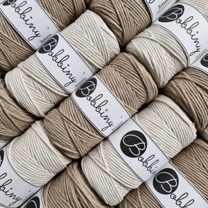 Peut inclure: Gros plan de plusieurs bobines de cordon en coton de 3 mm en couleurs naturelles et blanches. Les bobines sont étiquetées "Bobbiny" et présentent un logo en forme de lettre stylisée "B".