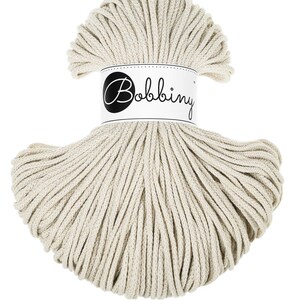 Può includere: Una matassa beige di cordoncino per macramè del marchio Bobbiny. Il cordoncino è avvolto strettamente e avvolto con un'etichetta bianca con il nome del marchio in nero, in corsivo. Il cordoncino è di colore neutro, adatto a vari progetti creativi.