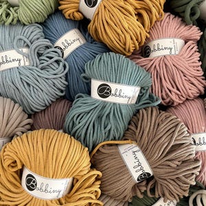 Può includere: Una varietà di colori di corda di cotone, tra cui verde, blu, marrone, rosa e giallo. La corda è disposta in una pila e ogni rotolo ha un'etichetta bianca con la parola "Bobbiny" stampata su di essa.