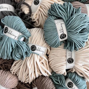 Puede incluir: Primer plano de varios carretes de cuerda de algodón en diferentes colores, incluyendo beige, azul verdoso y gris. Los carretes están etiquetados con "Bobbiny Premium 5mm Cotton Cord".