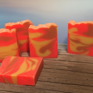 May include: Several bars of handmade soap with a vibrant orange, yellow, and brown swirl pattern. The soaps are rectangular with a textured top, and are arranged on a wooden surface. The background is a soft blue.