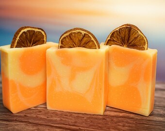 May include: Three square bars of handmade soap with orange and white swirls, each topped with a dried orange slice. The soaps are arranged on a wooden surface, with a blurred background of orange, blue, and purple.