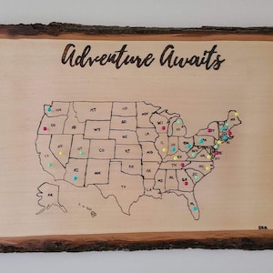 May include: A wooden map of the United States with the text "Adventure Awaits" at the top. The map is decorated with colorful push pins, indicating places visited.