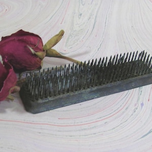 May include: A rectangular metal flower frog with numerous upright, sharp, dark-colored pins arranged in rows, positioned next to two dried, deep red roses with visible stems and withered petals, showcasing a contrast between the metal and organic elements.
