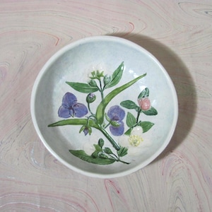 Puede incluir: Un cuenco redondo de cerámica blanquecina con un diseño floral. El cuenco presenta una ilustración botánica pintada a mano de flores moradas y blancas con hojas verdes. El cuenco está sobre un fondo veteado rosa y blanco.