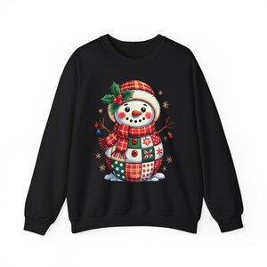 May include: Black sweatshirt featuring a whimsical snowman design. The snowman wears a plaid hat and scarf, with a patchwork body in red, green, and white. Decorative snowflakes and holly sprigs add to the festive holiday theme.