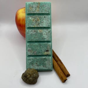 Op de afbeelding: Een rechthoekige, lichtgroene wax melt bar met zes secties, bespikkeld met bruin. Een rode en gele appel, een kleine, getextureerde steen en twee kaneelstokjes zijn rond de bar op een wit oppervlak geplaatst.