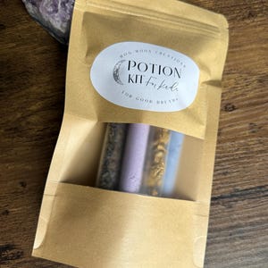 May include: A brown paper pouch with a clear window, holding four small glass vials filled with coloured contents. The pouch has a white oval sticker that reads "Potion Kit for Kids for Good Dreams". An amethyst crystal is visible in the background.