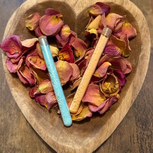 Puede incluir: Un cuenco de madera en forma de corazón lleno de pétalos de rosa secos en tonos rosa, rojo y amarillo. Dos tubos de vidrio, uno lleno de polvo azul y el otro de polvo naranja, descansan entre los pétalos. El cuenco está sobre una superficie de madera oscura.