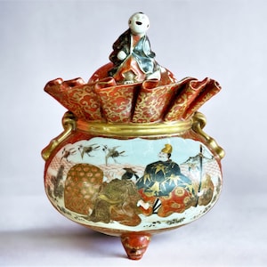 Puede incluir: Un quemador de incienso de cerámica rojo y dorado con una tapa que presenta una pequeña figura. El quemador está decorado con una escena de figuras y pájaros en un paisaje.