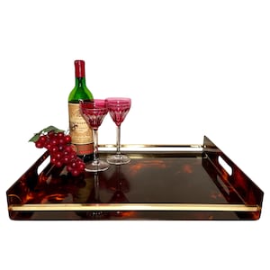 May include: A rectangular serving tray with a tortoiseshell pattern, gold trim, and handles. The tray holds a bottle of wine, two pink wine glasses, and a bunch of grapes. The tray is a decorative item.