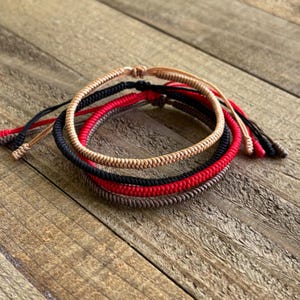 May include: A set of four braided cord bracelets in beige, black, red, and dark brown. Each bracelet features a round, woven texture with adjustable loose cord ends. The bracelets are stacked together, showcasing the distinct colours of the cords.