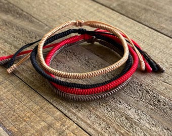 May include: A set of four braided cord bracelets in beige, black, red, and dark brown. Each bracelet features a round, woven texture with adjustable loose cord ends. The bracelets are stacked together, showcasing the distinct colors of the cords.
