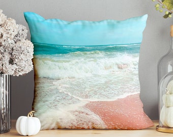 May include: A throw pillow featuring a beach scene with blue water, white waves, and pink sand. The pillow is on a wooden surface with a vase of flowers, a glass cloche with white pumpkins, and a white vase.