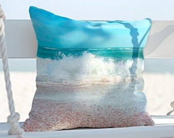 May include: A white swing with a blue and white cushion featuring a beach scene with waves crashing on the shore. The swing is hanging from a tree branch and is set against a blue sky and white sandy beach.
