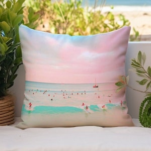 May include: A pink and blue throw pillow with a beach scene featuring surfers and a sailboat in the distance. The pillow has a white background and a soft, textured fabric.