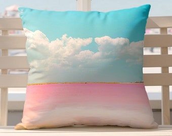 May include: A white cushion with a pink and blue landscape design. The cushion features a pink lake with a blue sky and white clouds. The cushion is sitting on a white wooden bench.